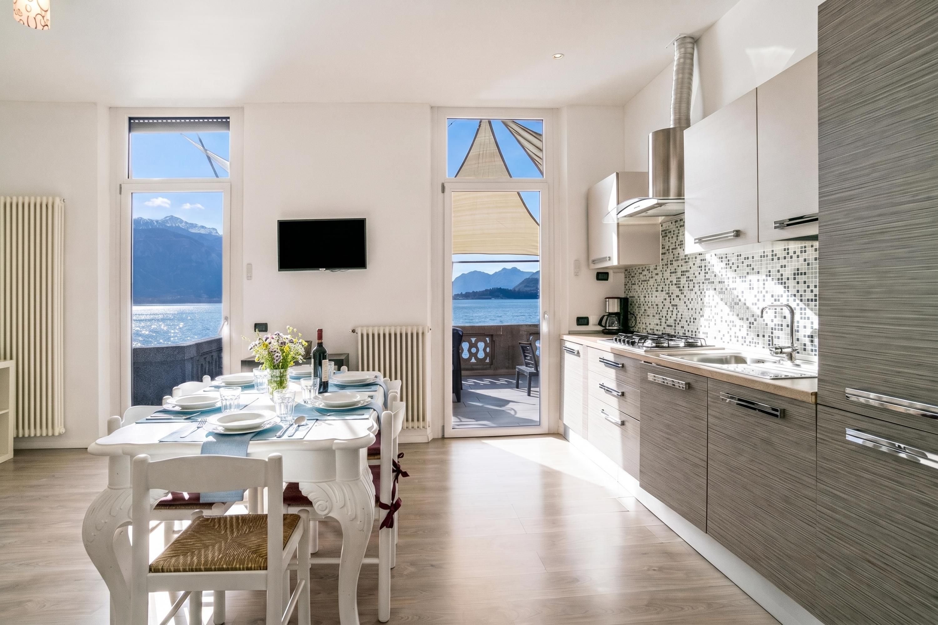 Una cocina moderna con vista al mar, un comedor con una mesa dispuesta para dos personas y un televisor montado en la pared.