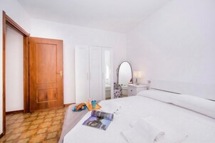 A hotel room with a wooden door, white wardrobe, a bed with white linens, a bedside table with a lamp, and a magazine on the bed.