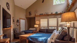 Uma sala de estar aconchegante com uma cama grande, um sofá, uma mesa de centro e uma televisão. Há duas janelas com persianas, um ventilador de teto e uma estante com objetos decorativos.