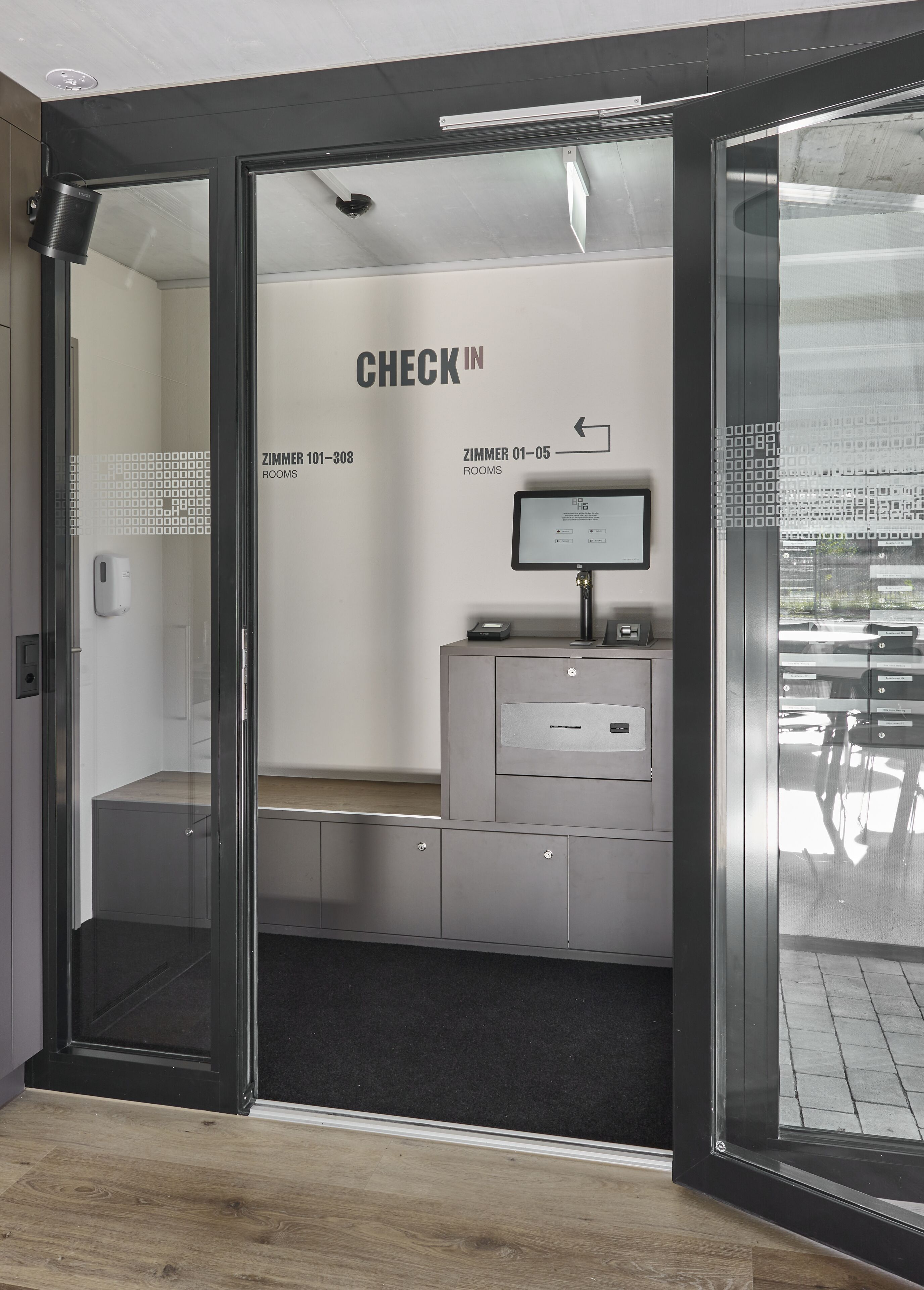 A check-in area with a monitor displaying room information, a reception desk, and a glass door leading to rooms.