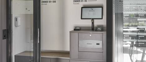 A check-in area with a monitor displaying room information, a reception desk, and a glass door leading to rooms.