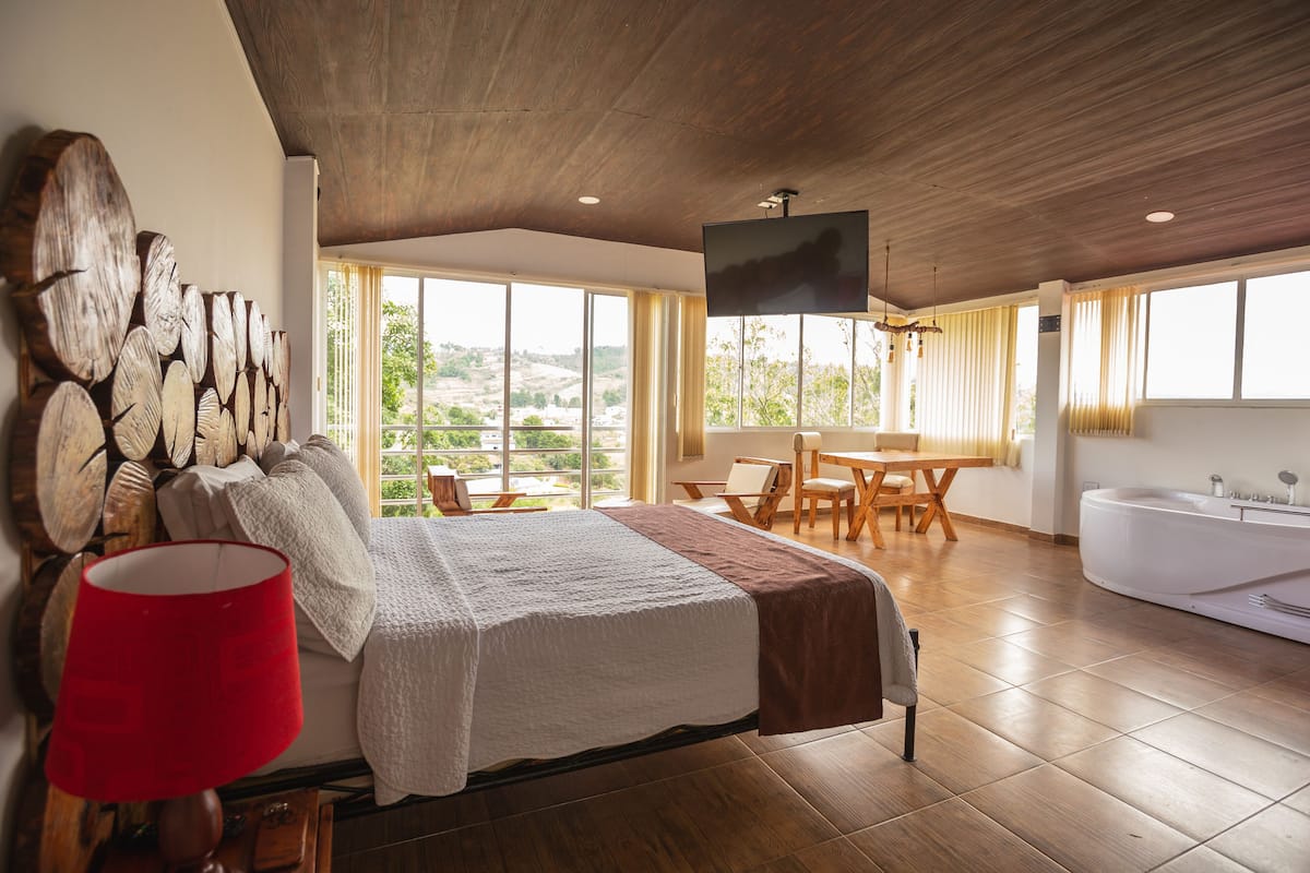 A bedroom with a large bed, a red lamp, a wooden wall art piece, a TV, a wooden table with chairs, and a view of the outdoors through large windows.
