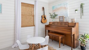 Una habitación con un piano de madera, una guitarra, una mesa redonda blanca con sillas y una ventana con persiana de bambú.
