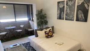 A bedroom with a bed, a small table, and a chair. There is a window with blinds, a plant, and artwork on the wall. - Barreiro Central Apartment  (Barreiro)