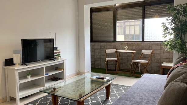 A modern living room with a flat-screen TV on a white TV stand, a glass coffee table, a sofa, and a dining area with two chairs and a small table. - Barreiro Central Apartment  (Barreiro)
