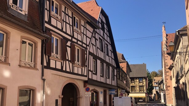 Une ruelle étroite pavée, bordée de bâtiments traditionnels à colombages, une voiture blanche garée sur le côté et un fourgon blanc au loin.