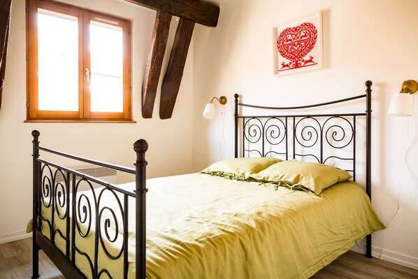 A bed with a metal frame and a green bedspread, a window with wooden shutters, and a wall-mounted light fixture. - Résidence des Tanneurs - Sylvaner (Obernai)