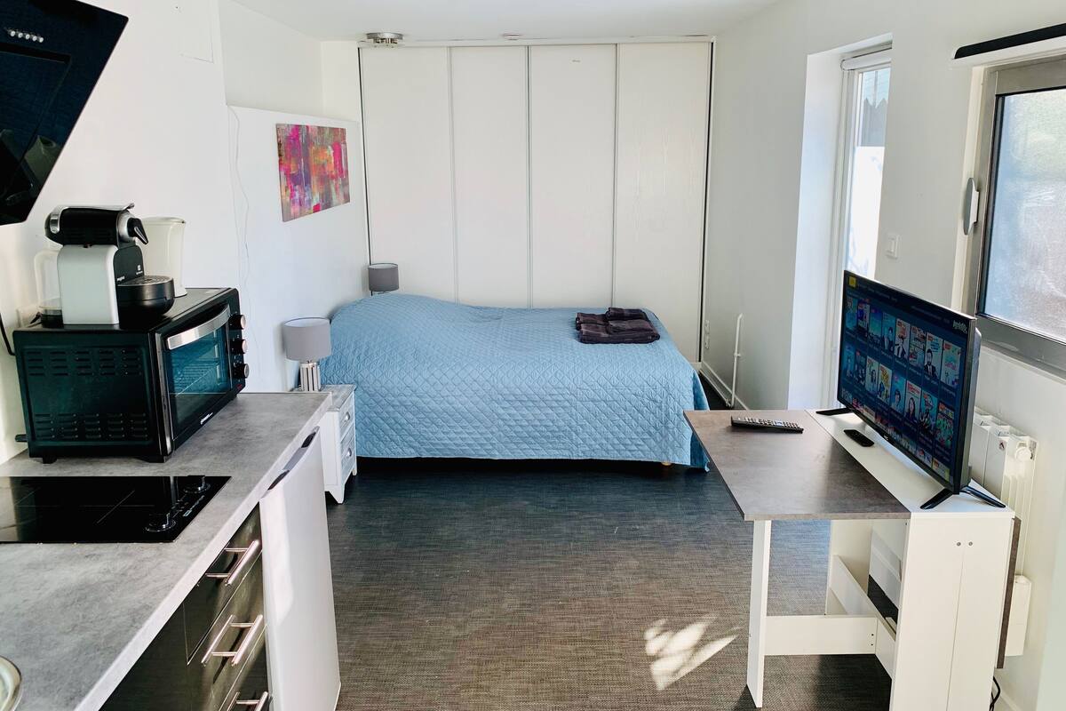 A compact hotel room with a kitchenette, a bed with a blue quilt, a TV, and a window.
