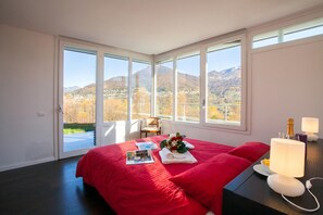 A bedroom with a large bed, a chair, a table with a lamp, and a view of mountains through large windows. - Villa La Betulla (Cerano d'Intelvi)