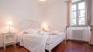 Une chambre à coucher avec un lit blanc, deux tables de chevet avec des lampes, un radiateur et une fenêtre avec des rideaux.