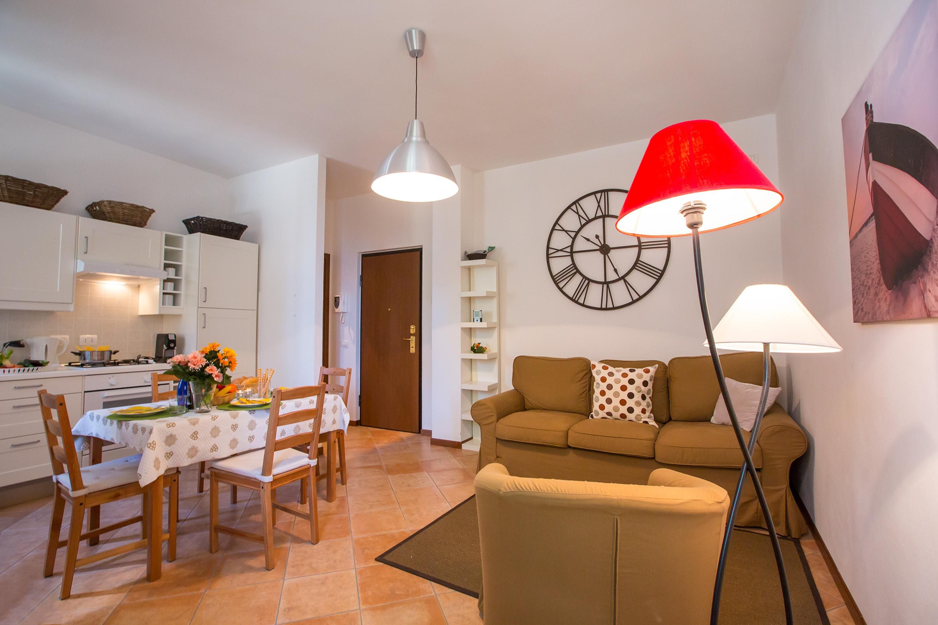 Een woonkamer met een bank, een eettafel met stoelen, een keukenhoek en een klok aan de muur.