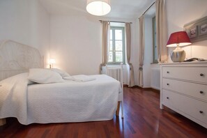 A bedroom with a bed, a dresser, a lamp, a window with curtains, and a radiator.