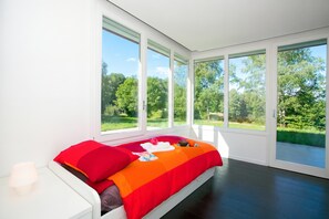 A bedroom with a large bed, white walls, and multiple windows offering a view of a green landscape. - Villa Intelvi (Cerano d'Intelvi)