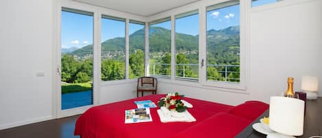 Un dormitorio con una colcha roja, una mesita de noche con lámpara y vistas a montañas y vegetación.