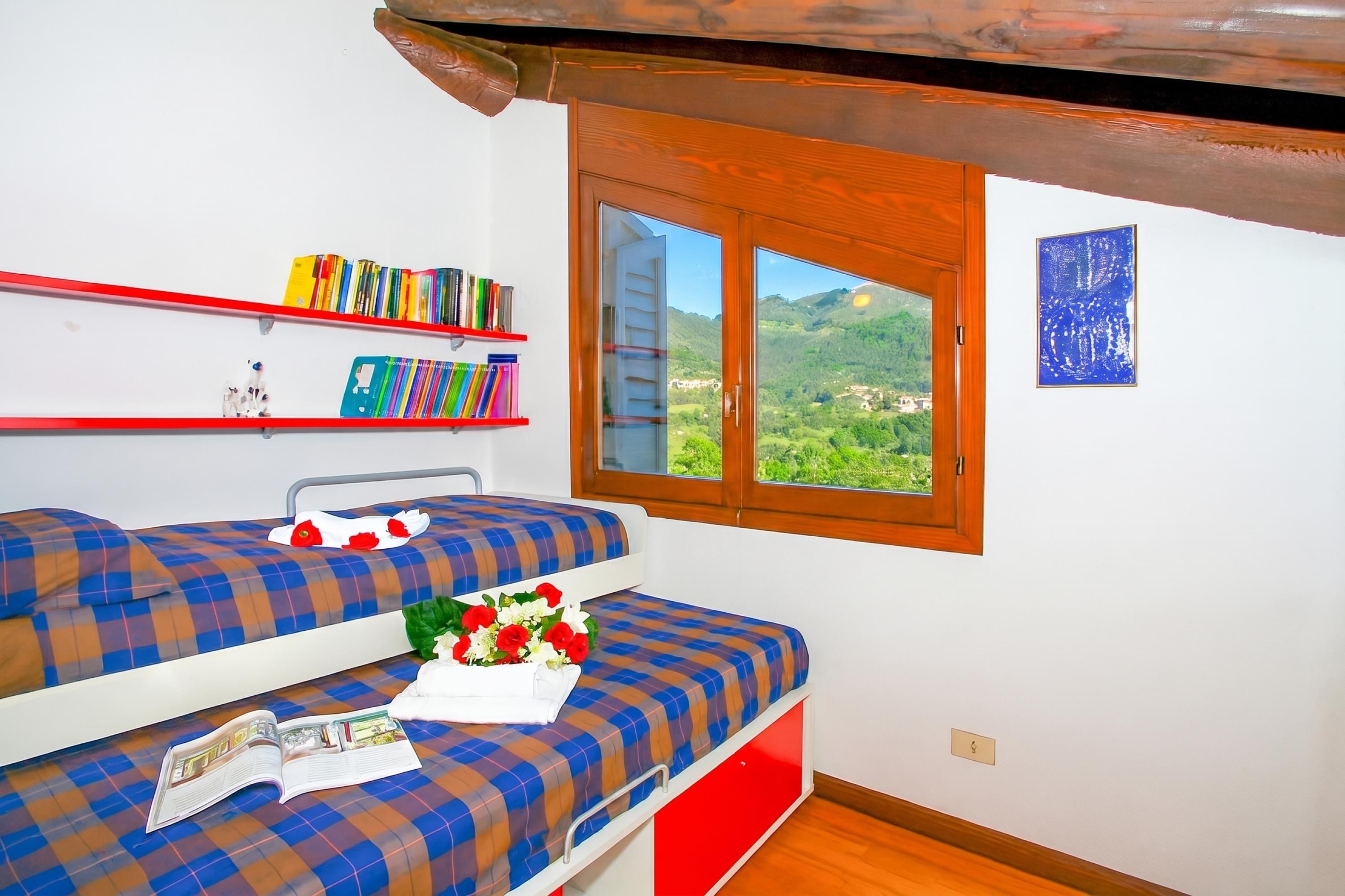 A room with two beds, a window with a mountain view, a bookshelf, and a framed picture on the wall.