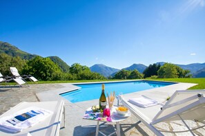 Escena junto a la piscina con sillas de descanso, una mesa con una botella, vasos y fruta, y montañas al fondo.