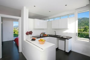 A modern kitchen with white cabinetry, a black countertop, and a view of a green landscape through large windows. - Villa Intelvi (Cerano d'Intelvi)