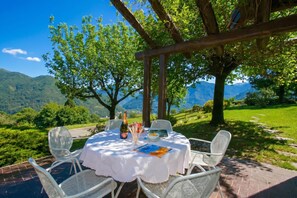 Un área de comedor al aire libre con una mesa dispuesta para dos personas, rodeada de sillas blancas, bajo una pérgola con vistas a un paisaje montañoso.