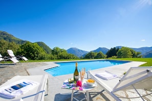 A poolside area with lounge chairs, a table with a bottle, glasses, and fruit, and a mountain view. - Villa Intelvi (Cerano d'Intelvi)