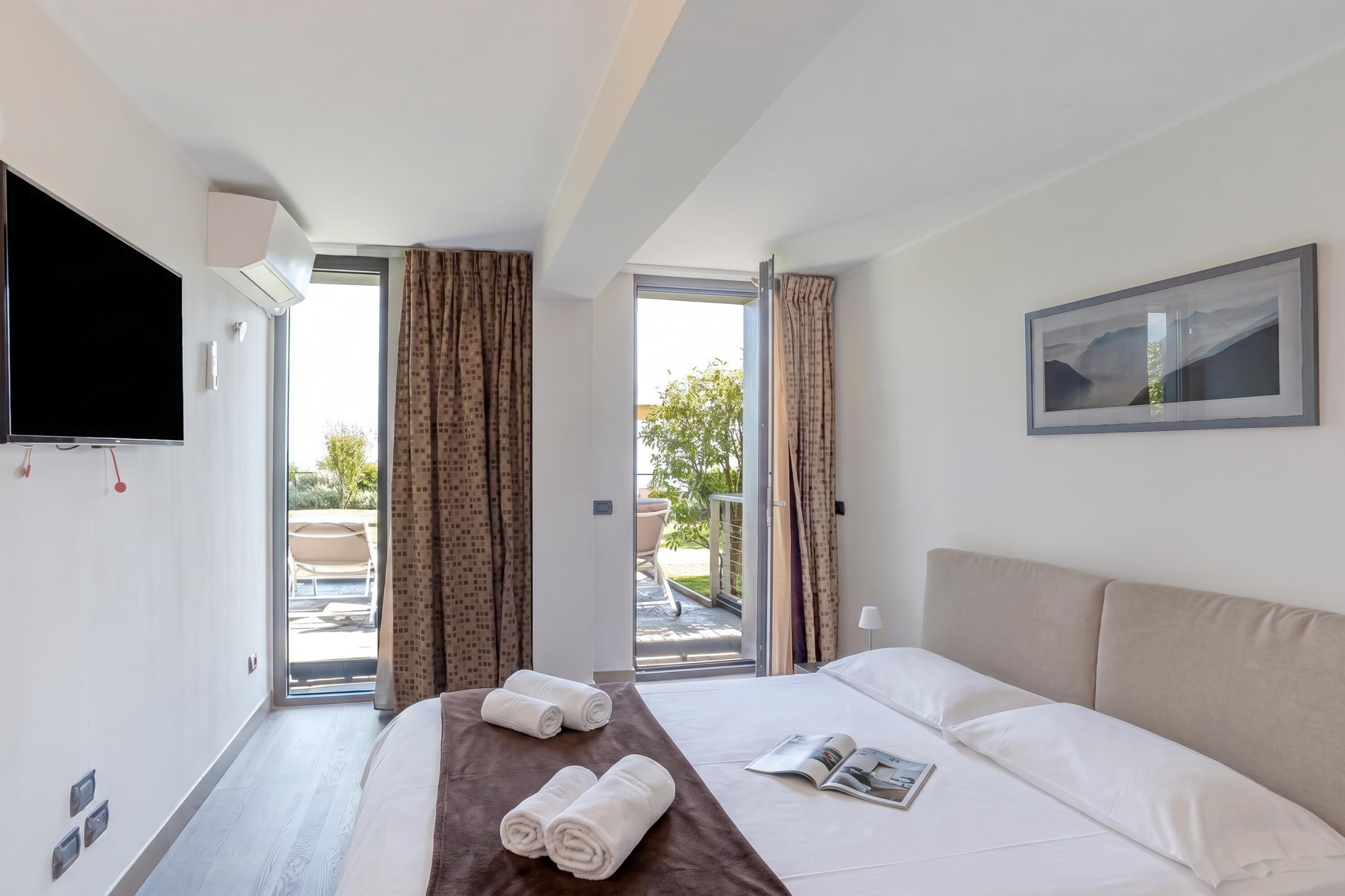 A hotel room with a bed, a flat-screen TV, a window with curtains, and a view of the outside.