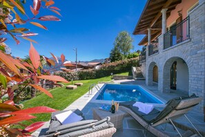 A pool area with lounge chairs, a table with a bottle and glasses, and a stone building with arched windows. - Villa Rosalia (Menaggio)