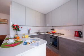A modern kitchen with white cabinets, a stainless steel sink, a red kettle, and a dining area with a green placemat.