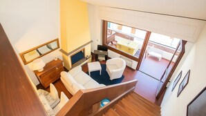 Un intérieur moderne avec un canapé, un fauteuil et une petite table, un escalier en bois et une vue sur un balcon extérieur.