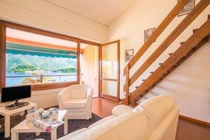 Une pièce comprenant un canapé, un fauteuil, une table avec un téléviseur et des théières, une fenêtre donnant sur un paysage de montagnes et d’eau, ainsi qu’un escalier en bois.