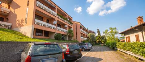 A multi-story residential building with balconies, parked cars, and a clear sky.