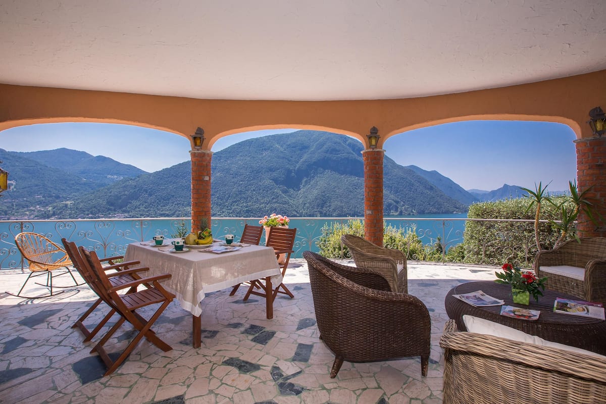 A terrace with a table and chairs, overlooking a mountainous landscape and a body of water.