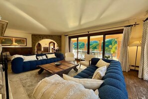 A living room with a blue sofa, a wooden coffee table, and a view of a lush garden through large windows. - Villa Isola Verde (Valsolda)