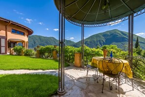A covered outdoor seating area with a table and chairs, a pathway leading to a house, and a mountainous landscape in the background. - Villa Isola Verde (Valsolda)