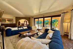 Un salon avec un canapé bleu, une table basse sur laquelle repose un livre, et une vue sur un paysage verdoyant grâce à de grandes fenêtres.