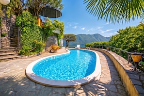 Une piscine dotée d’une terrasse en pierre, entourée d’une végétation luxuriante et offrant une vue sur les montagnes.