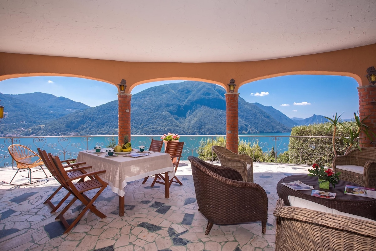 Una terraza con mesa lista para comer, mobiliario de mimbre y vistas a montañas y un lago.
