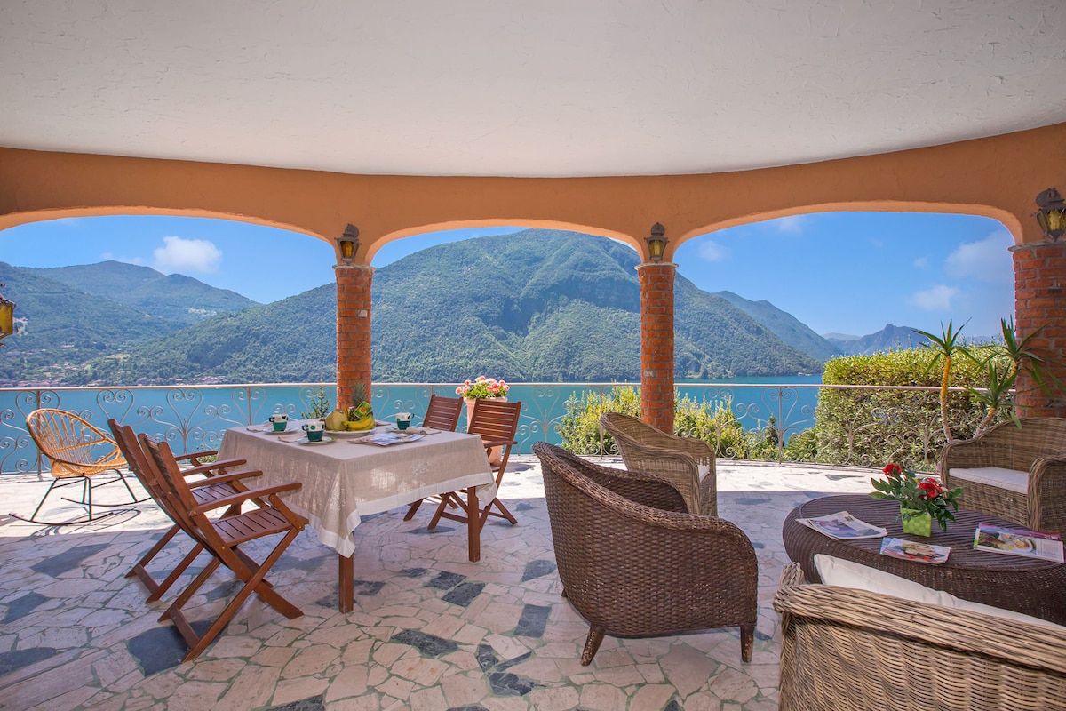 Une terrasse avec une table dressée pour le repas, des meubles en osier et une vue sur un lac et des montagnes.