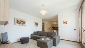 A modern living room with a grey sectional sofa, a small ottoman, a flat-screen TV, and a dining table with decorative vases. - Borgo La Sorgente Erica (Gravedona)