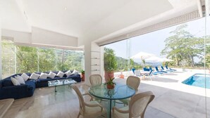 A spacious living area with a glass table and chairs, a blue sofa, and a view of a swimming pool and greenery. - Beautiful Villa with Pool and Bay Views in Acapulco (Acapulco de Juárez)
