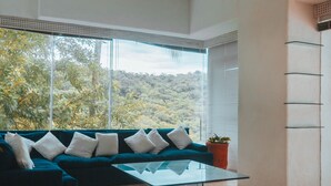 A modern living room with a teal sofa, a glass coffee table, and a large window offering a view of a forested area. - Beautiful Villa with Pool and Bay Views in Acapulco (Acapulco de Juárez)