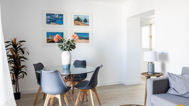 Spiseområde med et glasbord, fire stole, en vase med blomster og billeder i rammer på væggen.