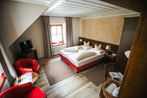 A hotel room with a bed, red chairs, a TV, a window, and a wooden floor. - Landhotel Lindenhof (Feld am See)