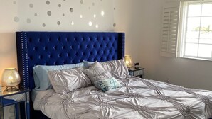 Ein Schlafzimmer mit einem samtenen, blauen Kopfteil, einem großen Bett mit floralem Bettbezug, zwei Nachttischlampen und einem Deckenventilator.