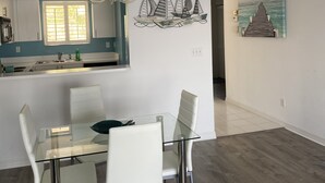Ein moderner Essbereich mit einem Glastisch und weißen Stühlen, einer Kristallleuchter sowie einer Küche mit Fenster und einem Gemälde eines Piers an der Wand.