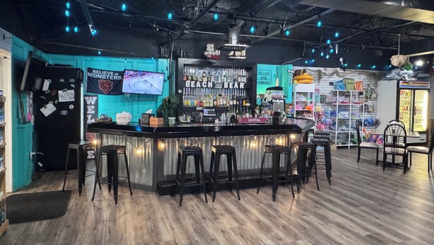 Zona de bar con mostrador, taburetes, una pantalla de televisión y una estantería con diversos objetos.