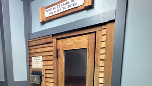 Una puerta de madera con un cartel que indica restricción de edad, un letrero que advierte contra verter agua sobre rocas o equipos eléctricos, y un picaporte de metal.
