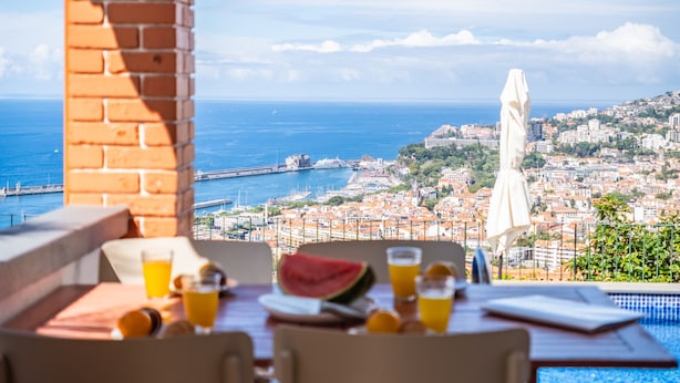 Una mesa con bebidas y una sandía, con vistas a una ciudad costera y al mar, donde se aprecian edificios.