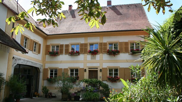 Ein zweistöckiges Gebäude mit einer symmetrischen Fassade, zahlreichen Fenstern mit Fensterläden und Blumenkästen im oberen Bereich.