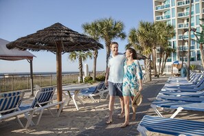 A beachside hotel with lounge chairs, a thatched umbrella, and palm trees. - MONTHLY RENTAL - Gorgeous Modern Ocean View Condo w/ Access to Resort Amenities (Myrtle Beach)
