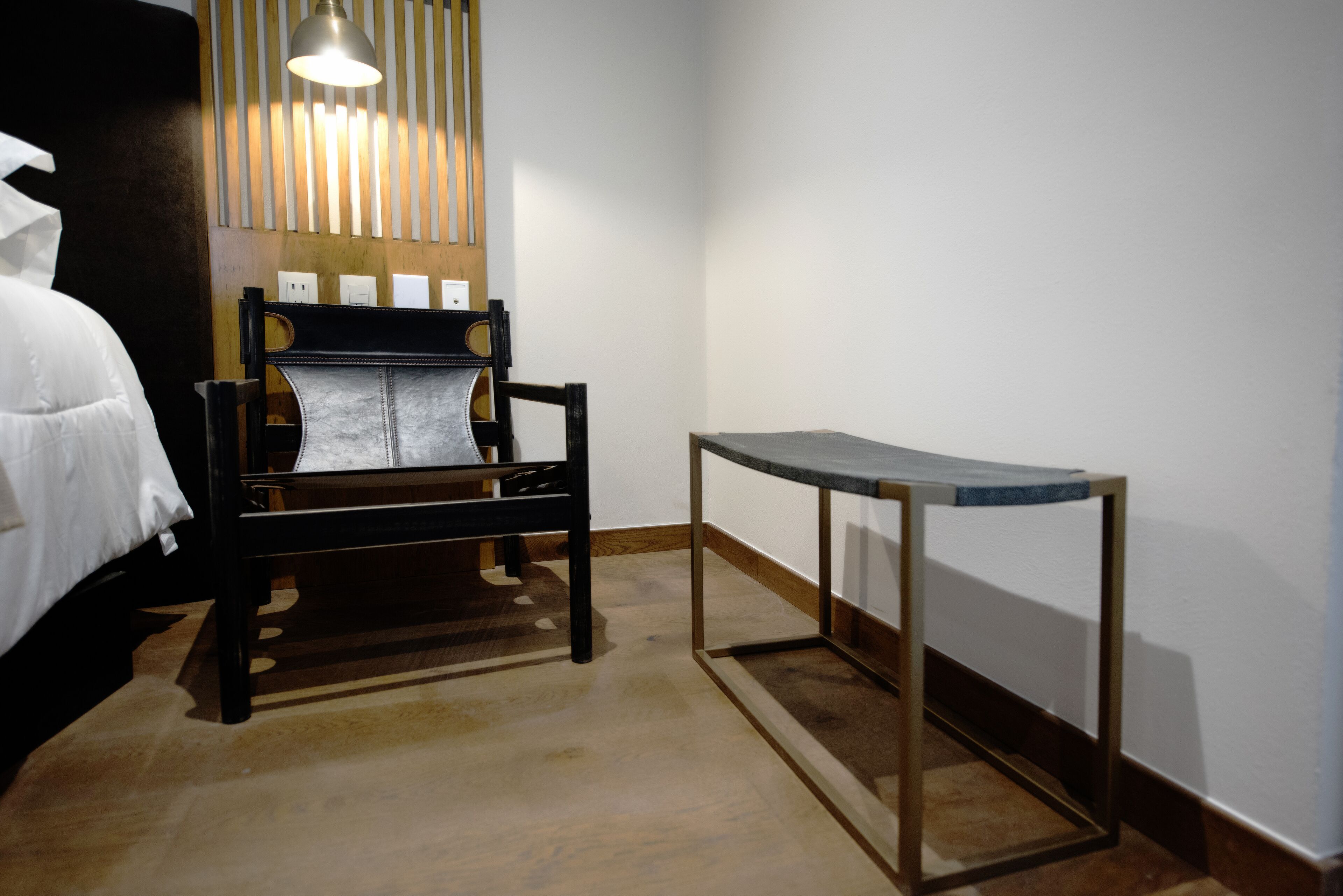 A hotel room with a bed, a wooden chair, a small table, and a wall-mounted light fixture.