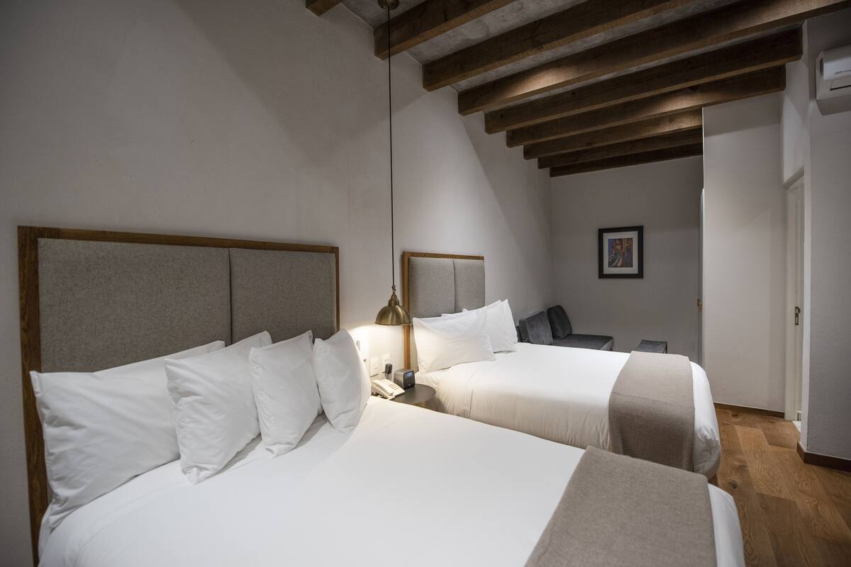 Habitació d'hotel amb dos llits, una taula de nit amb una làmpada, un quadre al mur i bigues de fusta al sostre.